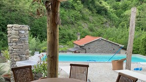 Patio esterno in legno con tavolo e sedie rotonde, un pilastro in pietra e vista su una piscina e un edificio in pietra sullo sfondo.