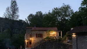 Una casa in pietra con una scala in pietra che conduce a un portico coperto. È presente un braciere attorno al quale sono sedute persone.