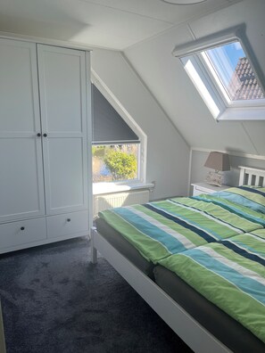 Ein Schlafzimmer mit schrägem Dach, einem Oberlicht, einem Bett mit gestreifter Bettdecke, einem Nachttisch mit Lampe und einem Kleiderschrank.
