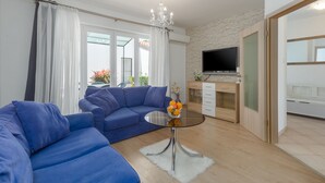 A modern living room with a blue sectional sofa, a glass coffee table with fruit, a chandelier, and a TV mounted on a stone-textured wall. - Family-friendly, quiet location, near the beach, barbecue in the garden, (Poreč)