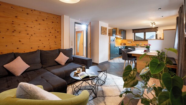 Ein modernes Wohnzimmer mit Sofa, Couchtisch und Sesseln, ein Küchenbereich mit Esstisch und Blick auf Berge durch das Fenster.