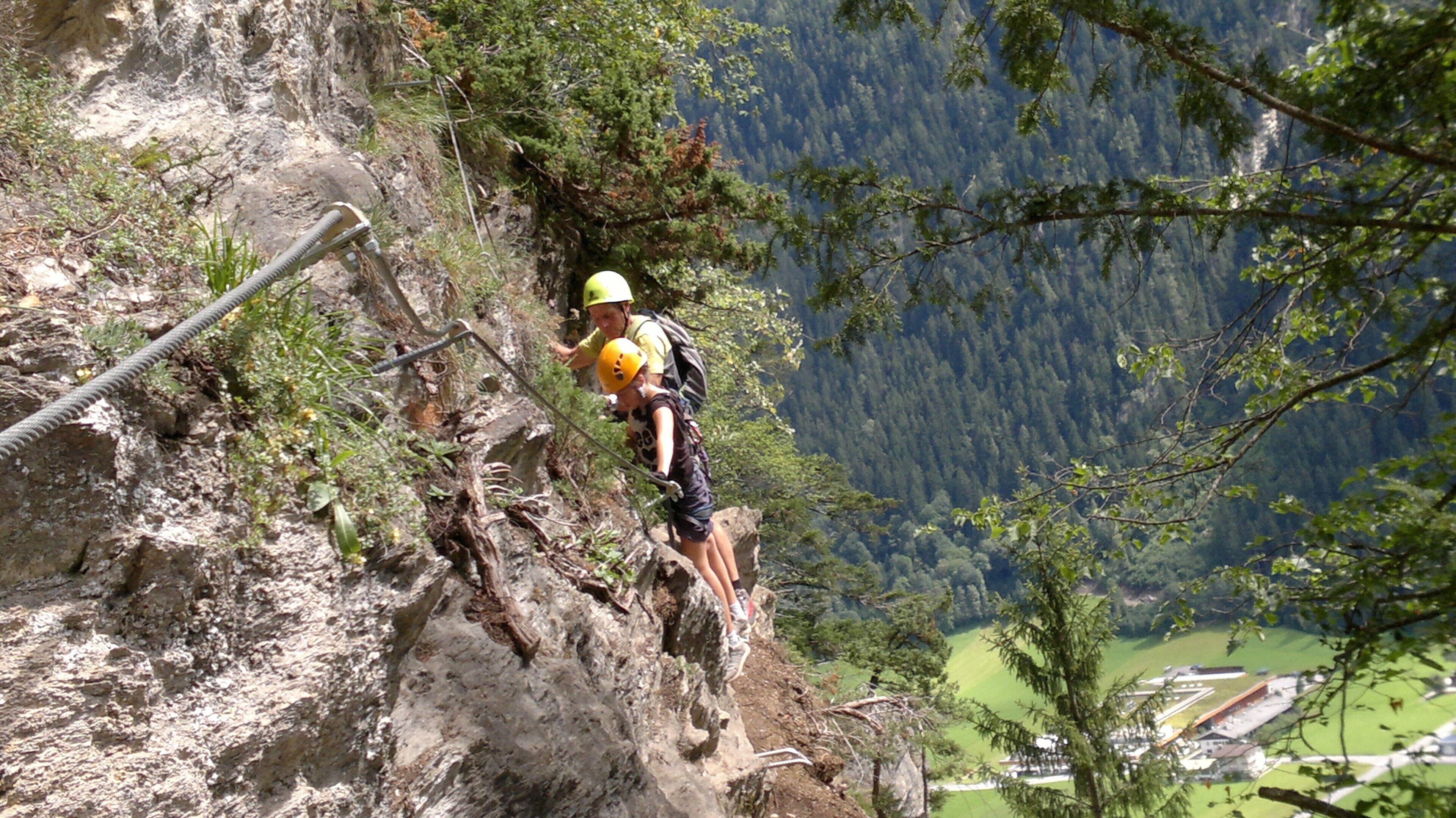 Eine Person klettert auf einem steinigen Bergpfad, im Hintergrund ein bewaldetes Tal.