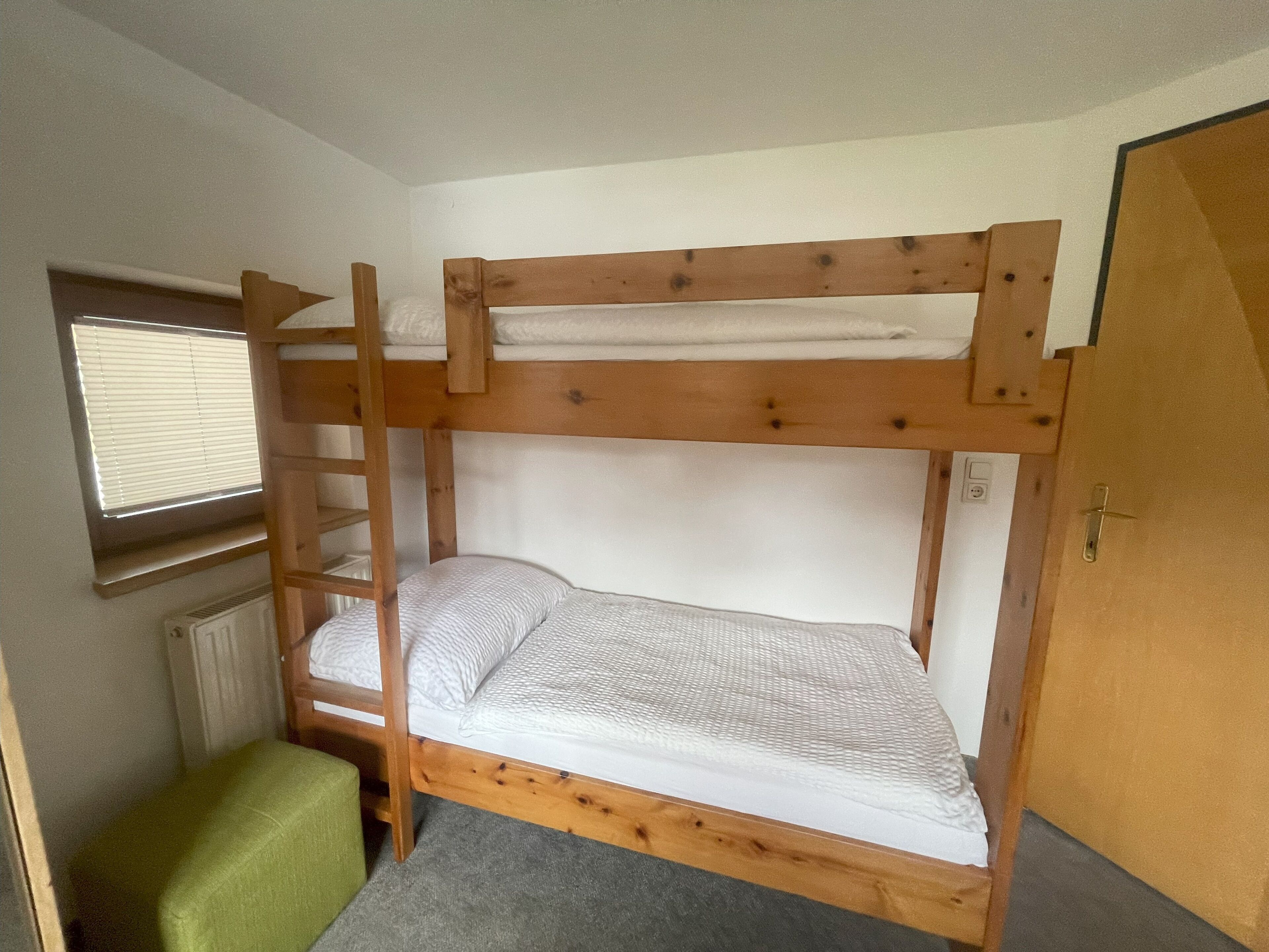 Ein Stockbett mit einer Leiter, ein kleiner grüner Stuhl und ein Fenster mit Jalousien.