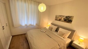 Et pænt møbleret soveværelse med en stor seng, et sengebord med lampe, et vindue med gardiner og et billede hængt på væggen.