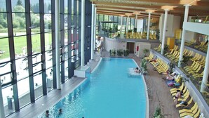 En indendørs swimmingpool med flere baner, liggestole og store vinduer med udsigt over en anlagt have.