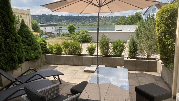 A patio with outdoor furniture, a large umbrella, and a view of a lake and distant buildings. - Modern, premium lake view vacation apartment over 2 floors (Waldeck -Scheid)