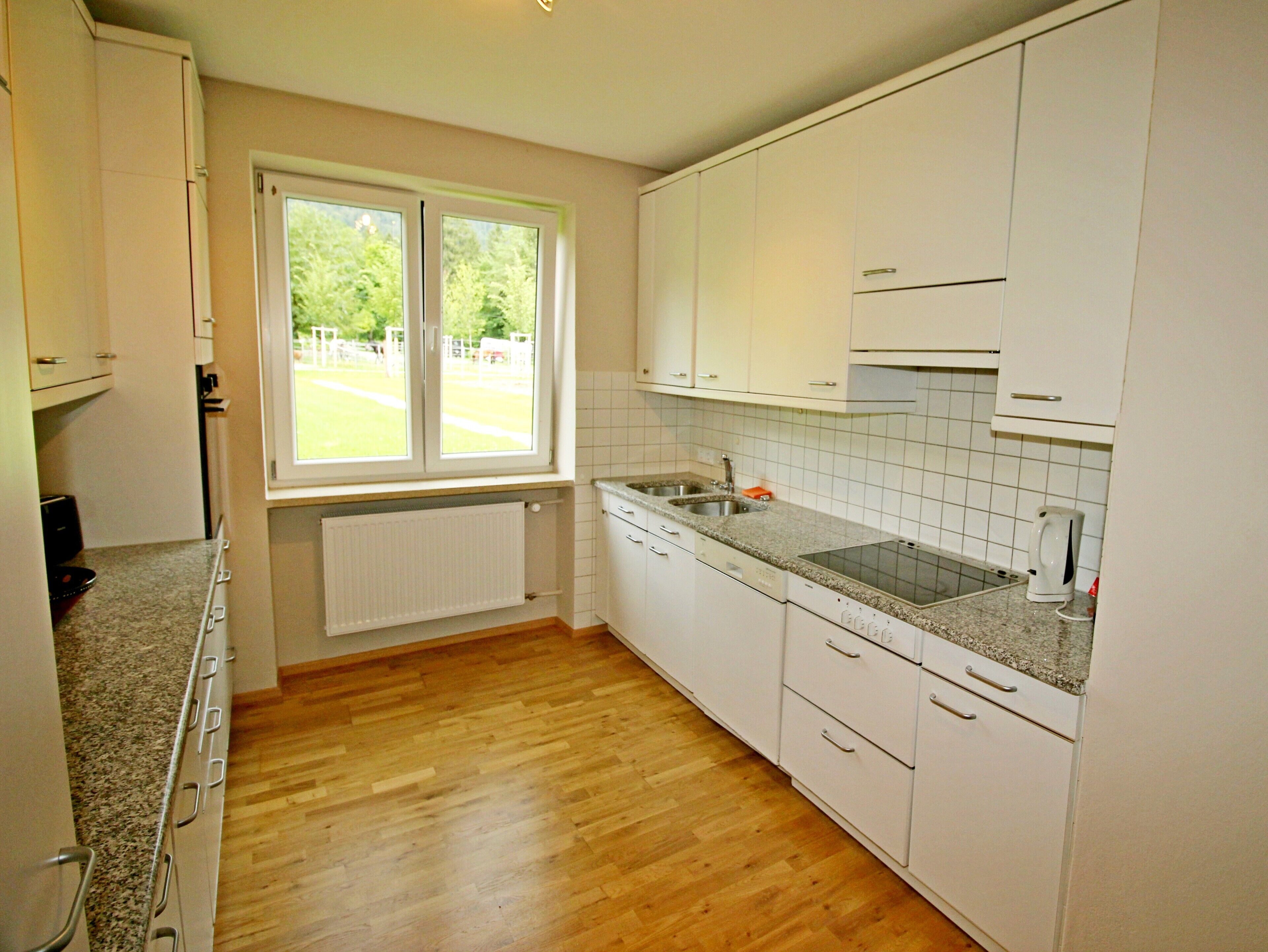 A kitchen with white cabinets, a granite countertop, a double sink, and a window with a view of greenery outside.