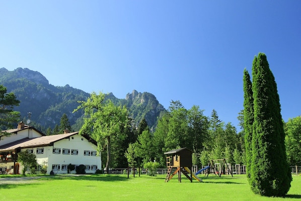 A playground with a slide and swings, surrounded by green grass and trees, with a mountain in the background.