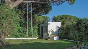 A small, white building with a door and window, surrounded by lush greenery and a tree.