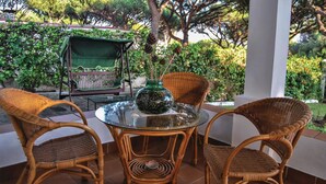 A glass-top table with wicker chairs, a green vase with pinecones, and a bench with a green canopy in a garden setting.