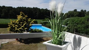 Een witte plantenpot met een plant op een balkon, met een zwembad op de achtergrond.