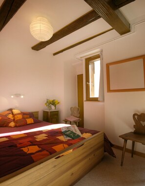A bedroom with a wooden bed, a small table with a vase of flowers, a chair, and a window with a wooden frame.