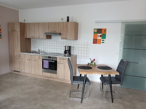 A modern kitchen with wooden cabinets, a built-in oven, a coffee maker, and a dining area with two chairs.