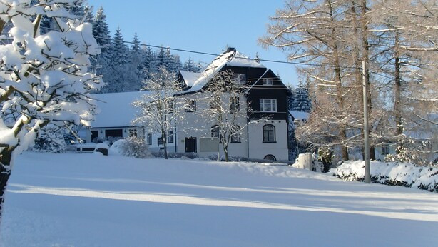 Een huis bedekt met sneeuw, met een zwart dak en witte muren, omgeven door sneeuwzware bomen en een heldere lucht.
