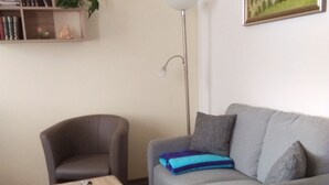 Een woonkamer met een grijze bank, een kleine houten tafel, een vloerlamp en een boekenkast met boeken en een plant.