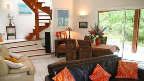 Een woonkamer met een trap, een bank, een eethoek met rieten stoelen, en uitzicht op groen buiten.
