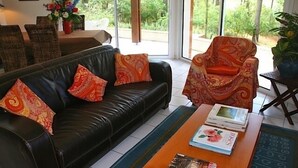 Een woonkamer met een zwarte leren bank, gekleurde kussens, een houten salontafel met boeken, en uitzicht op groen door grote ramen.