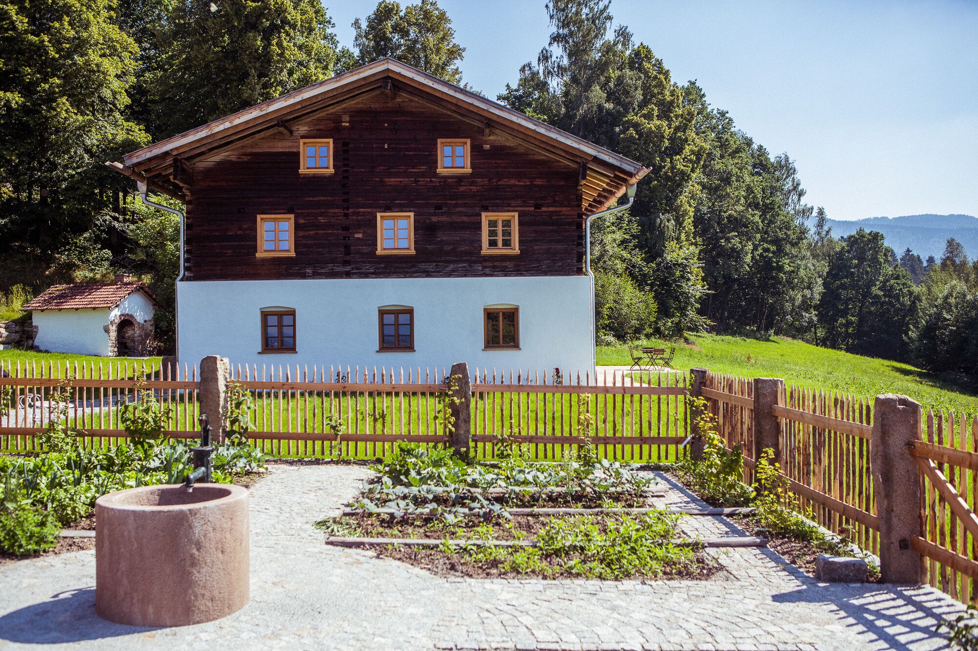 A two-story wooden house with a white lower section and a dark upper section, surrounded by a wooden fence and a garden path.