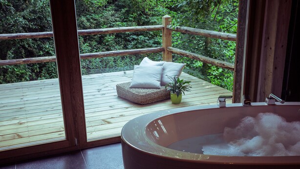 A wooden deck with a hot tub, a cushioned seating area, and a potted plant.