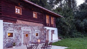 A rustic wooden house with stone walls, a stone entrance, and a patio with a table and chairs.