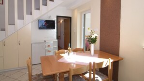 Un coin repas avec une table et des chaises en bois, un petit réfrigérateur, un téléviseur fixé au mur et un escalier menant à une autre pièce.
