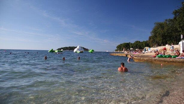 Une plage à l’eau claire, où l’on peut voir des gens nager et des bouées gonflables vertes flotter.