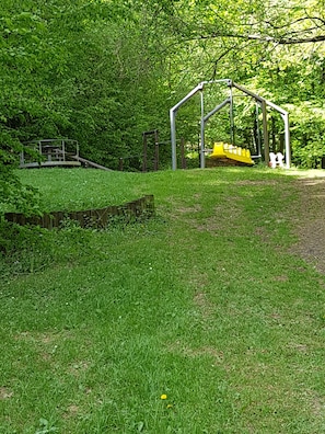 Children can play safely in this enclosed outdoor area surrounded by lush greenery, featuring a slide and a swing set.