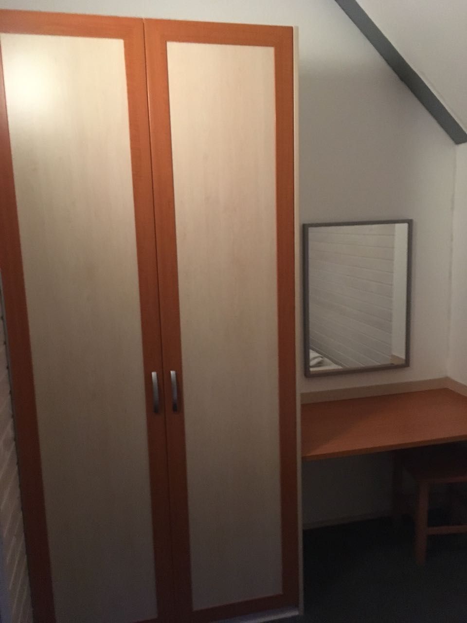 A room with a wardrobe, a desk, and a mirror on the wall.