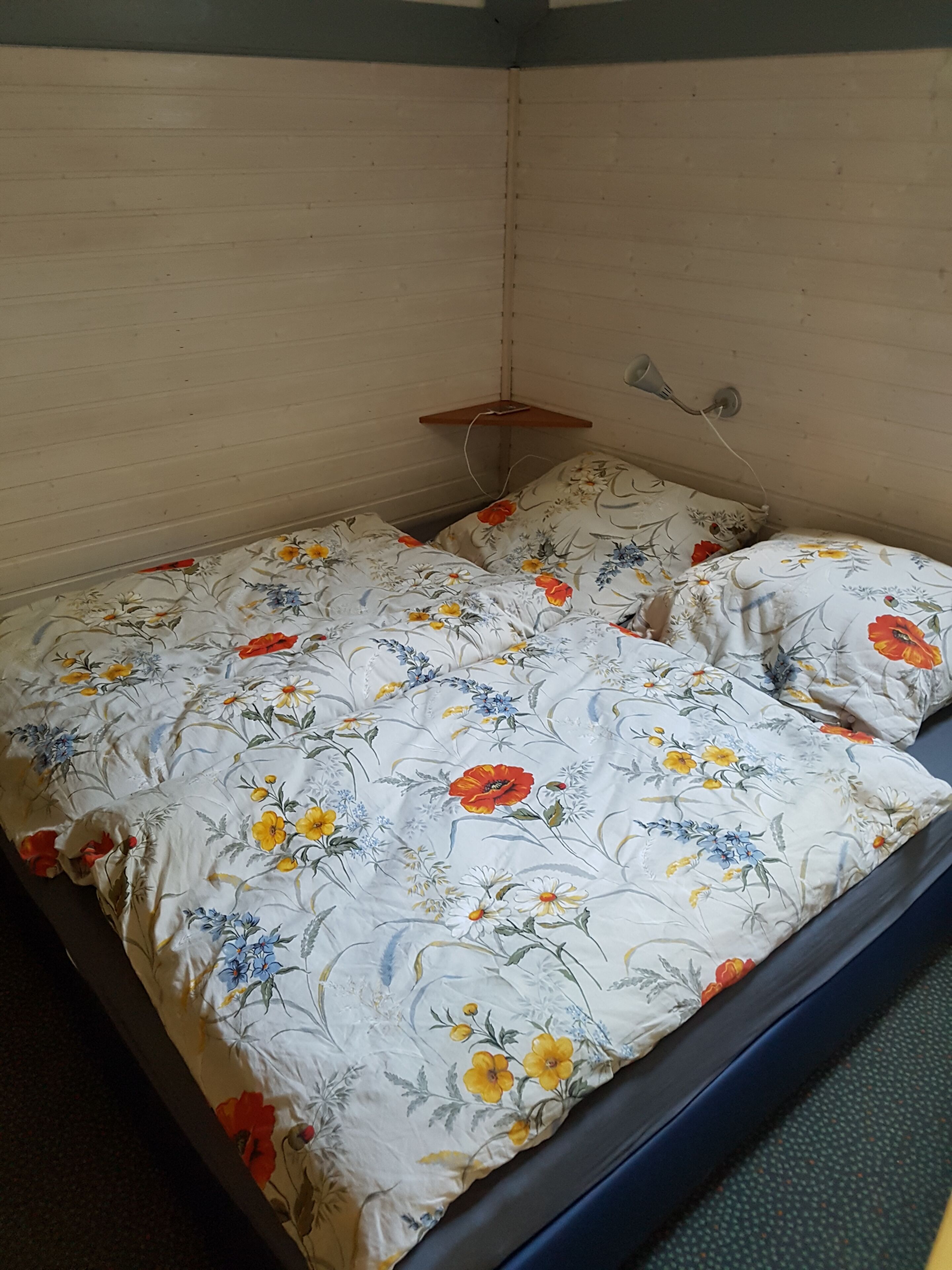 A small, single-bed room with a floral-patterned bedspread and a wooden shelf.