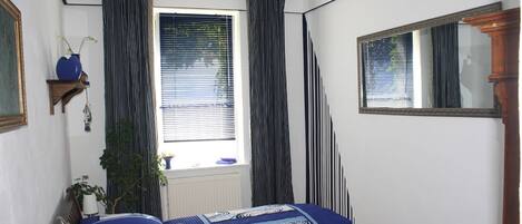 Ein Schlafzimmer mit Bett, Vorhängen am Fenster, einem Spiegel und einer Pflanze.
