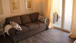 A living room with a brown sofa, two patterned pillows, a white throw blanket, a floor lamp, and a sliding glass door leading to an outdoor area with potted plants.