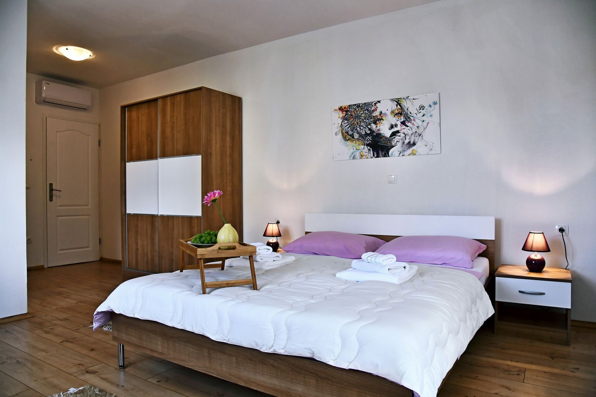A bedroom with a bed, a bedside table, a nightstand, a wall-mounted air conditioner, a door, and a painting on the wall.