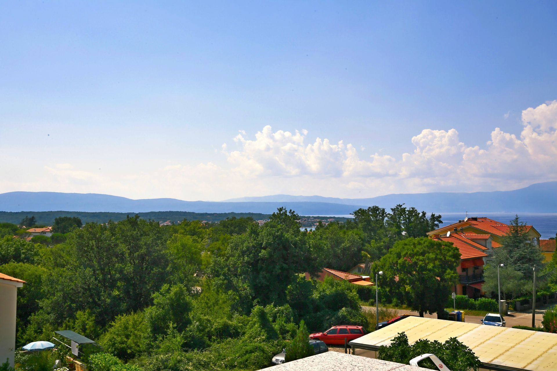 A residential area with houses, a red car, and a view of the sea and mountains.