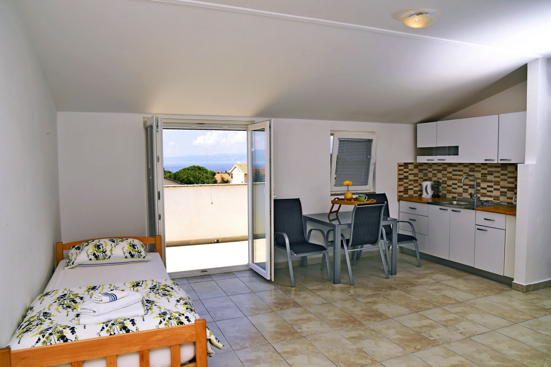A small, well-lit room with a bed, a dining table with chairs, a kitchenette, and a sliding glass door leading to a balcony with a view of the sea and buildings.