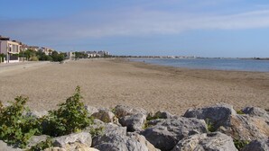 A sandy beach with rocky outcrops in the foreground, a clear sky, and buildings in the background. - Very nice vacation home, top location, only 50 m to the beach, close to the harbor. 6 persons (Gruissan)
