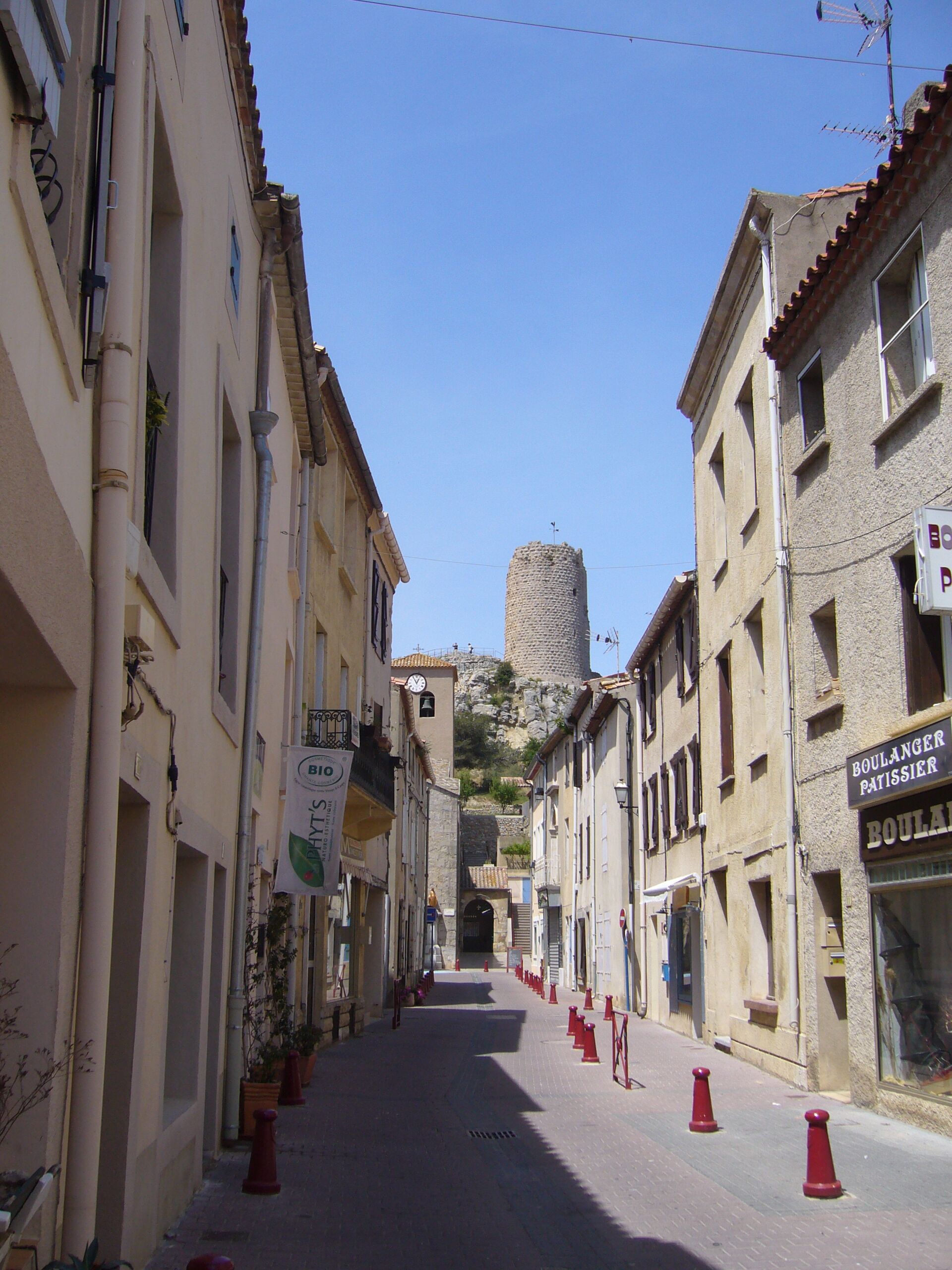 Eine schmale Straße mit Häusern an beiden Seiten, ein Glockenturm in der Ferne und ein Schild für eine Boulanger Patisserie.