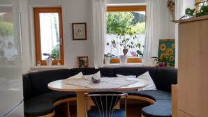 Een gezellige woonkamer met een donkergrijze bank, een ronde houten tafel en een stoel. Er zijn twee ramen met gordijnen, een potplant en een ingelijst beeld aan de muur.