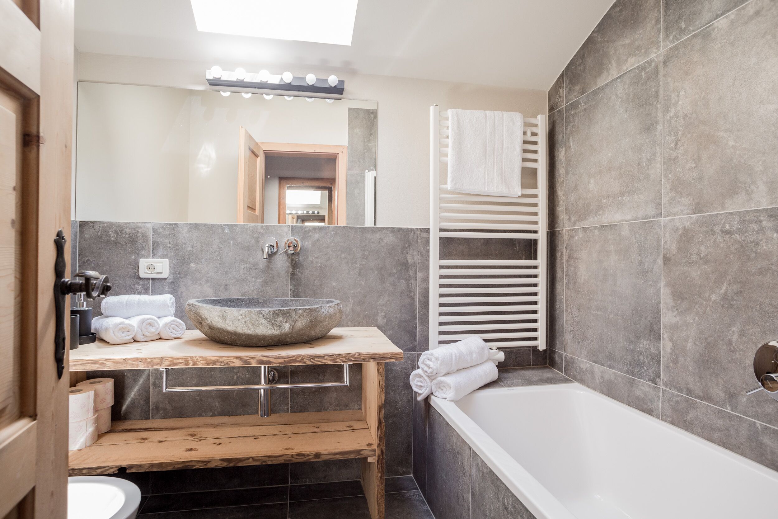 A modern bathroom with a stone sink, wooden vanity, a bathtub, and a mirror with lights.