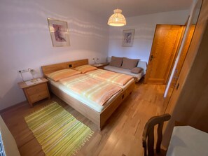 A bedroom with a wooden bed, bedside tables, a nightstand, a hanging lamp, a sofa, a door, and a chair.
