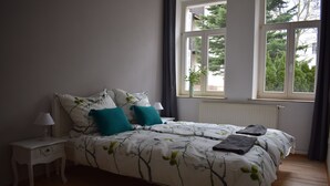 A bedroom with a bed, a nightstand, and a window with a view of trees. - Apartment, close to the center and in a quiet location on the floodplain forest (Leipzig)