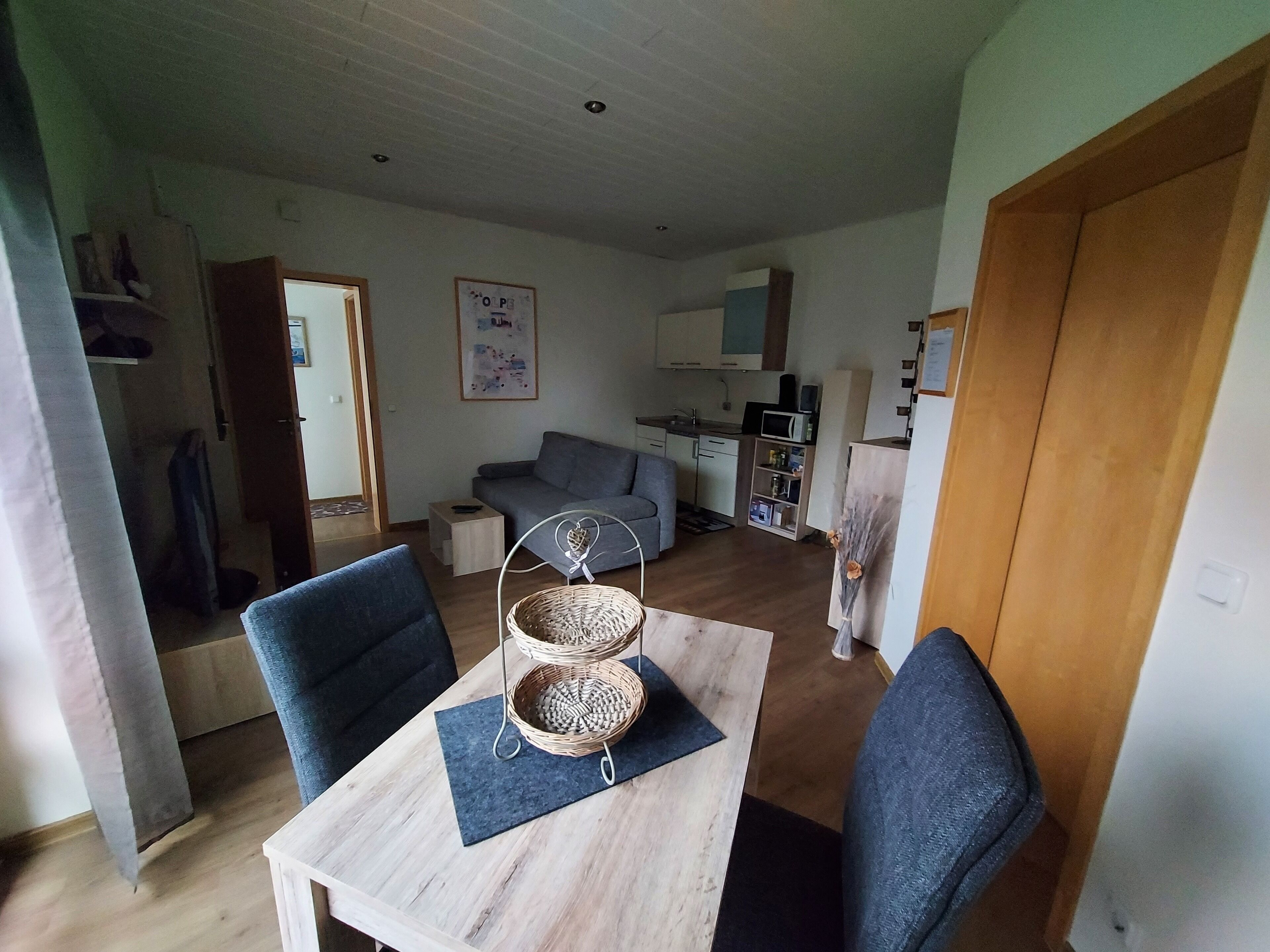 A dining area with a table, chairs, and a decorative basket. There is a sofa and a kitchen in the background.
