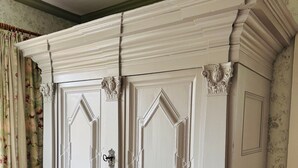A white, ornate armoire with decorative moldings and a lock, positioned in a room with patterned wallpaper and wooden flooring.