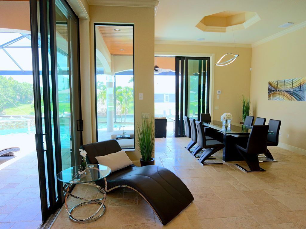 Une chambre d’hôtel moderne dotée d’une porte vitrée donnant sur un balcon, d’un canapé en cuir noir, d’une table basse en verre, d’une table à manger avec des chaises et d’une grande fenêtre offrant une vue sur l’extérieur.