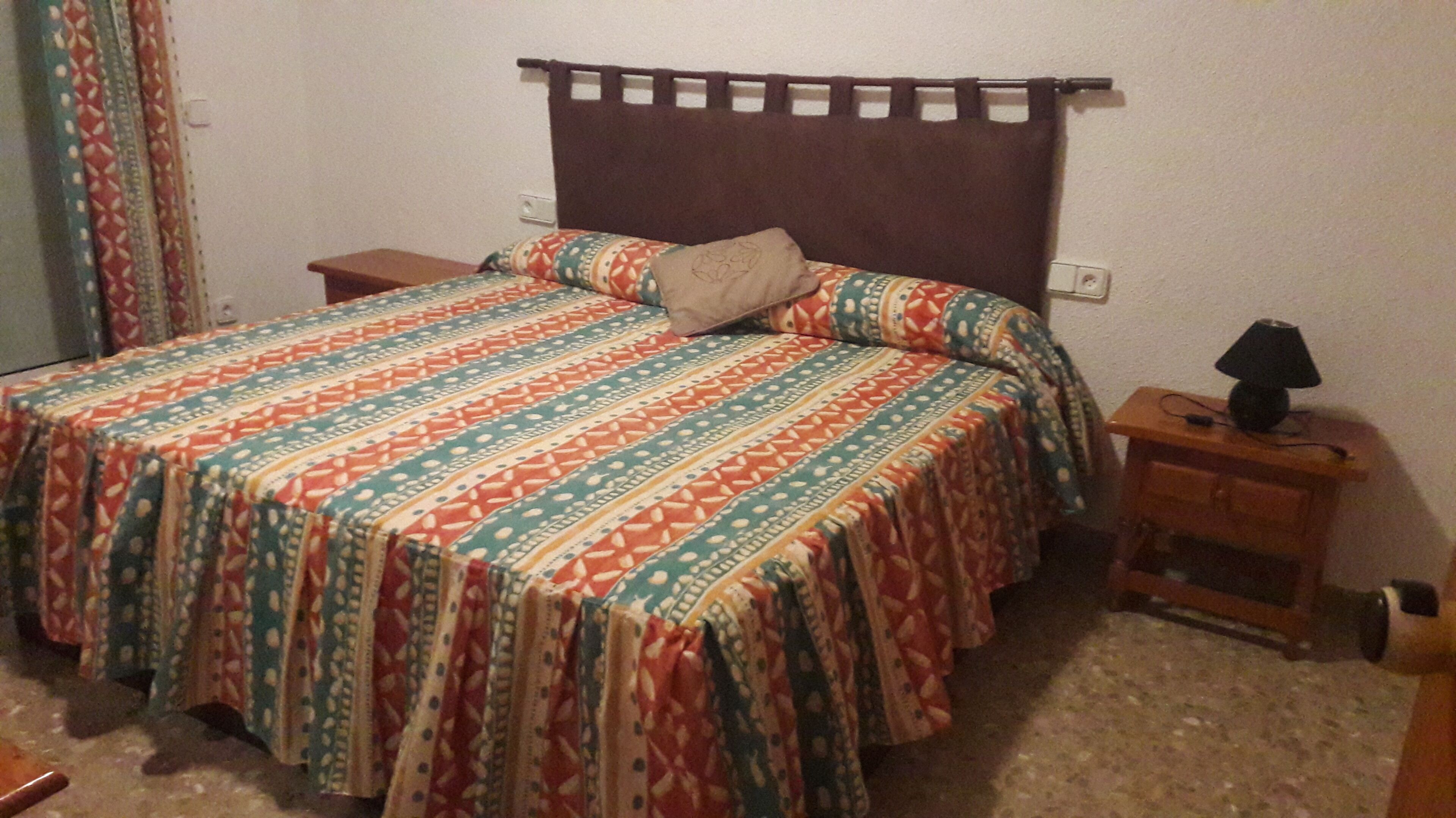A bed with a patterned bedspread, a bedside table with a lamp, and a power outlet on the wall.