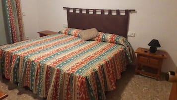 A bed with a patterned bedspread, a bedside table with a lamp, and a power outlet on the wall.