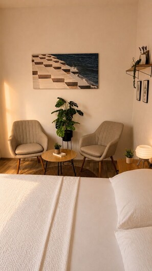 Ett hotellrum med en säng, två fåtöljer, ett litet bord, en krukväxt, en tavla som föreställer en strand och en hylla med dekorativa föremål.
