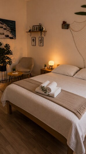 A bedroom with a bed, a chair, a nightstand with a lamp, a plant, and a painting on the wall.