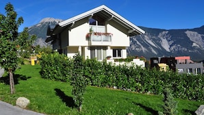 Une maison à deux étages dotée d’un balcon, entourée d’une végétation luxuriante et avec des montagnes en toile de fond.