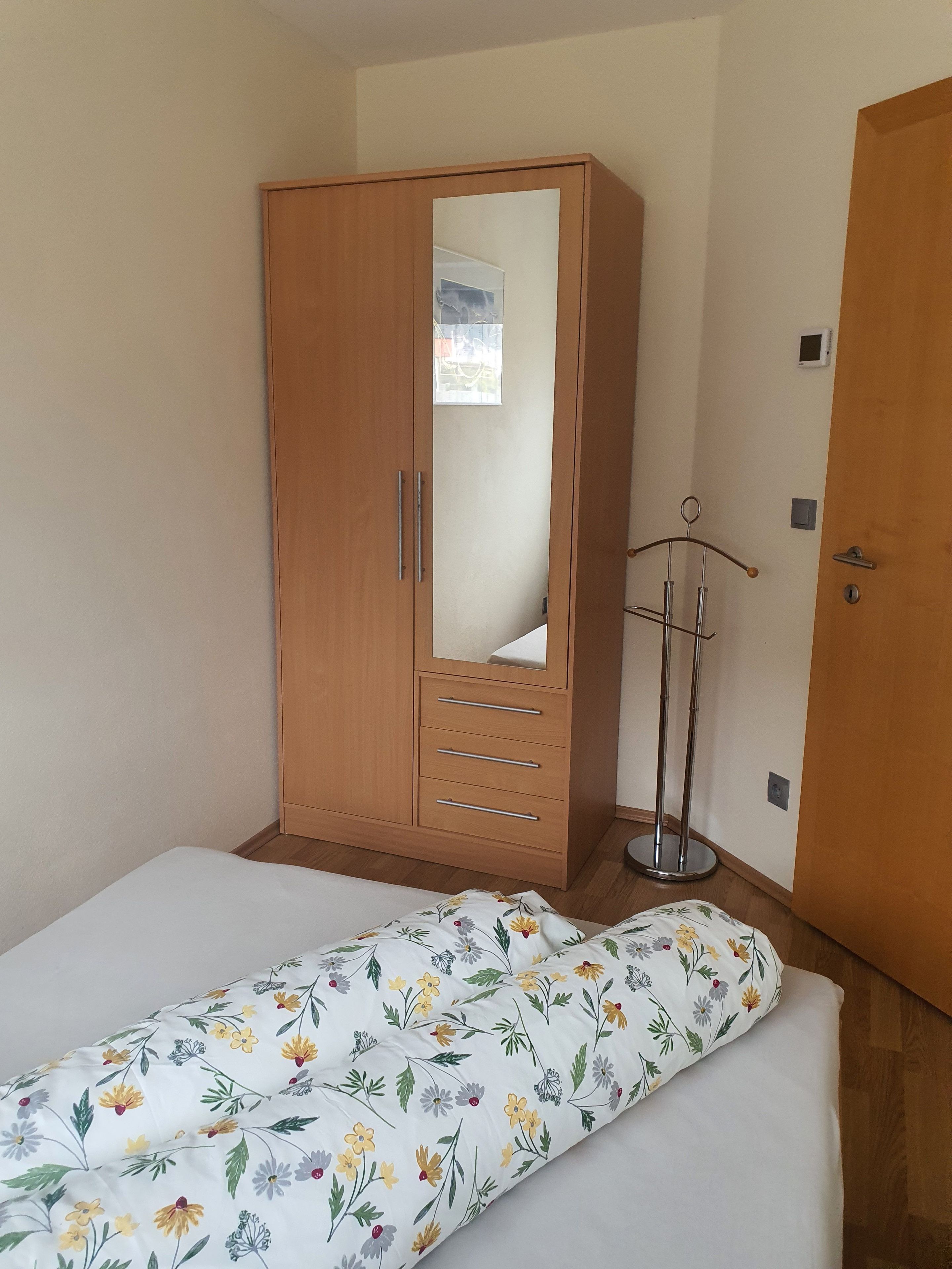 A bedroom with a wooden wardrobe, a bed with floral bedding, and a bedside table with a lamp.