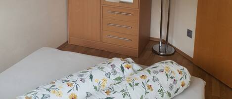 A bedroom with a wooden wardrobe, a bed with floral bedding, and a bedside table with a lamp.